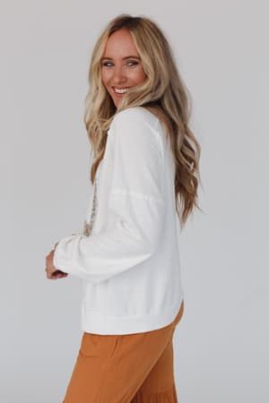 A woman with long, wavy blonde hair smiles over her shoulder, wearing the Jenny Floral Peace Sign Pullover Top in off white and mustard-colored pants, standing against a plain light background.