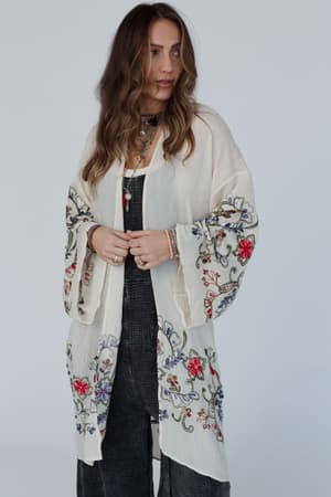A woman with wavy brown hair wears the Fun In Florals Kimono - Cream, featuring delicate floral embroidery over a dark outfit. She accessorizes with layered necklaces and bracelets, standing against a plain light background.