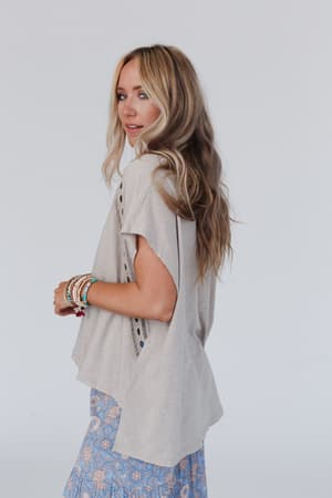 A woman with long wavy blonde hair wears the Camden Lace V Neck Top in Camel and a blue floral skirt, accessorized with beaded bracelets, as she stands against a light background, looking over her shoulder.