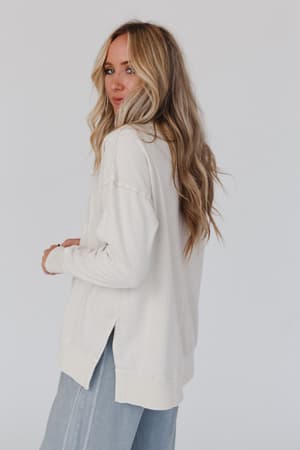 A woman with long wavy blonde hair models the Catch My Smile Pullover Top in khaki and light blue pants, standing sideways and looking over her shoulder against a plain white background.