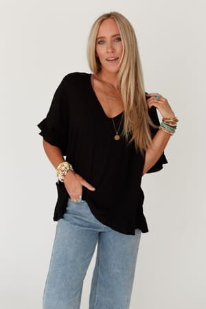A woman with long blonde hair poses against a white background wearing The Nest Let Me Be Ruffled Top in black, paired with light blue jeans and accessorized with multiple bracelets and rings.