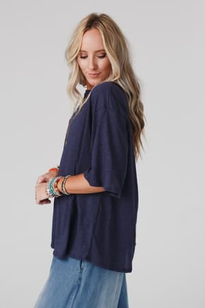 A woman with long, wavy blonde hair wears The Nest Your Favorite Slub Tee Shirt in navy and light blue jeans, standing sideways and looking down. She accessorizes with several colorful bracelets against a plain light background.