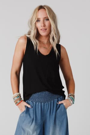 A woman with wavy blonde hair wears The Nest Shelli Scoop Neck Tank Top in black, loose blue pants, layered necklaces, and colorful beaded bracelets. She stands with hands in her pockets against a plain light background.
