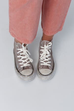 A person wearing light pink pants and the Chantel Studded High Top Sneaker in Mocha stands on a white surface.