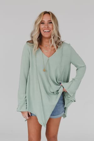 A woman with wavy blonde hair smiles in the Golden Hour Long Bell Sleeve Top - Sage and denim shorts, layering gold necklaces, standing against a plain light background.