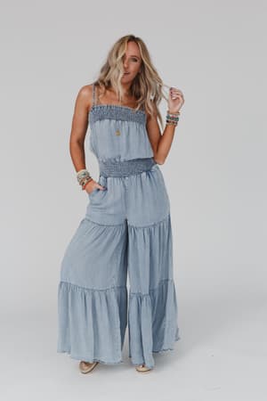 A woman with wavy blonde hair poses against a white background, wearing the Harlow Wide Leg Chambray Jumpsuit - Blue, styled with chic bracelets and sandals.