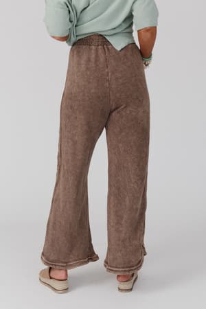 A person stands facing away, wearing the So Comfy Wide Leg Pant Full Length in Mocha with a light green top and beige sandals against a plain white background.