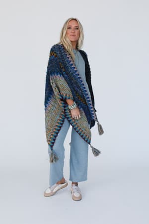 A woman with long blonde hair wears a light blue jumpsuit and the Desert Dreams Crochet Ruana - Cobalt, its colorful knit and tassels adding a boho vibe as she stands confidently against a white background.
