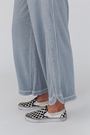 A person wearing the So Comfy Wide Leg Pant Cropped Length in Faded Blue with a high-rise elastic waist, small side slit at the hem, and black-and-white checkered slip-on shoes stands on a white background.