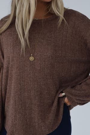 A person with long blonde hair wears The Nest Sweet Heart Balloon Sleeve Knit Sweater in Mocha and a gold circular pendant necklace, with one hand on their hip. Their face is not visible.