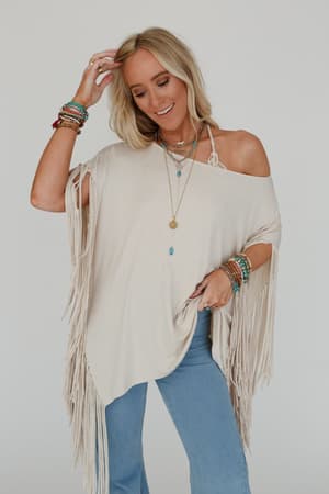 Woman wearing a beige fringe poncho and blue pants against a white background