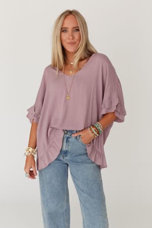 A woman with long blonde hair wears The Nest Let Me Be Ruffled Top in Pale Plum and blue jeans, accessorized with layered necklaces, bracelets, and rings, standing against a plain white background.