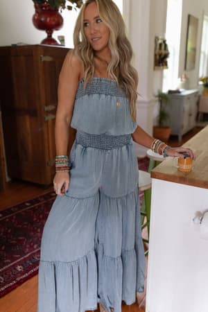 A woman with long blonde hair stands by a kitchen counter holding a mug, wearing the Harlow Wide Leg Chambray Jumpsuit - Blue and colorful bracelets. The room has wooden furniture, plants, and a rug in the background.
