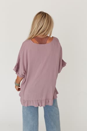 A woman with long blonde hair faces away, wearing The Nest Let Me Be Ruffled Top in Pale Plum layered over a brown bralette, paired with light blue jeans against a plain white background.