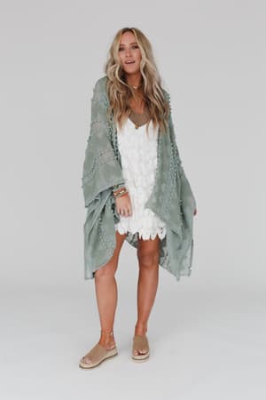 A woman with long, wavy blonde hair wears the Bali Embroidered Kimono in Sage over a white scalloped dress. She pairs it with tan sandals and colorful bracelets on her left wrist, standing against a plain background.