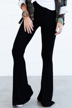 Wearing the Cher Solid Raw Edge Flare Pant in black, a white top, a camouflage jacket, and layered bracelets, the person stands on a light floor against a white brick wall. The photo captures them from shoulders to feet.