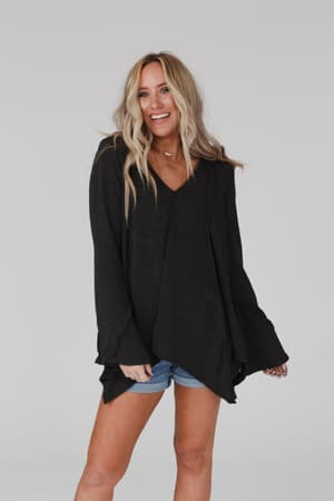 A woman with long, wavy blonde hair smiles while wearing the Golden Hour Long Bell Sleeve Top in Charcoal and denim shorts against a plain light gray background.