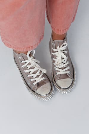 A person wearing light pink pants and Chantel Studded High Top Sneakers in mocha, featuring white laces and silver studs on the soles, stands on a light-colored surface.