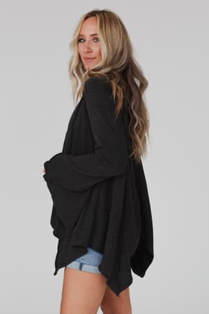 A woman with long blonde hair wears the Golden Hour Long Bell Sleeve Top in Charcoal and denim shorts, standing sideways, smiling over her shoulder against a plain, light background.