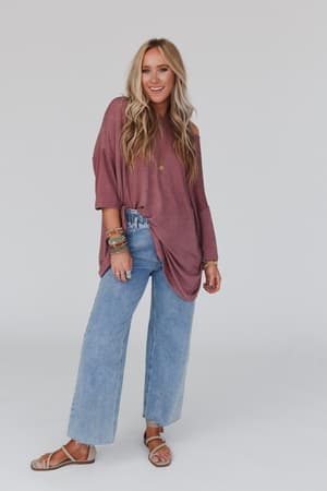 A woman with long blonde hair styled in soft waves smiles against a plain background, wearing The Nest Sunday Stroll Oversized Tee in mauve, paired with light blue wide-leg jeans, tan sandals, and colorful bracelets.