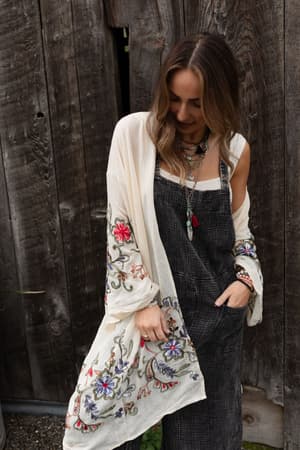 A woman stands before a rustic wooden wall, wearing the Fun In Florals Kimono - Cream over a dark dress. The sheer kimono features colorful floral embroidery. She accessorizes with layered necklaces and bracelets and smiles softly, her hair in loose waves.