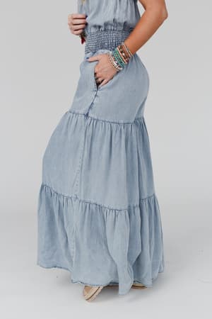 A woman wearing the Harlow Wide Leg Chambray Jumpsuit in blue, styled with colorful beaded bracelets and light shoes, stands against a plain background, exuding a relaxed boho vibe with her hand in her pocket.
