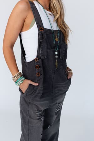 Wearing the High Hopes Slouchy Pocket Denim Overalls with black pockets and a white tank top, she accessorizes with layered turquoise and gold jewelry. Her loose, wavy hair adds to her effortlessly laid-back style.