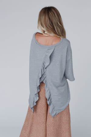 A woman faces away, wearing The Nest Must Have High Low Tee in heather gray, featuring a ruffled back seam in soft stretch knit. She pairs it with a flowing light brown patterned skirt; her blonde hair is down against a light gray background.