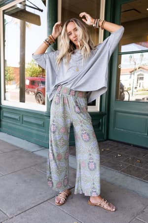A woman stands outside a shop in The Nest Cozy Cool Oversized Tee - Heather Gray, patterned wide-leg pants, and studded sandals. She poses with her hand in her hair, accessorized with bracelets and layered necklaces.