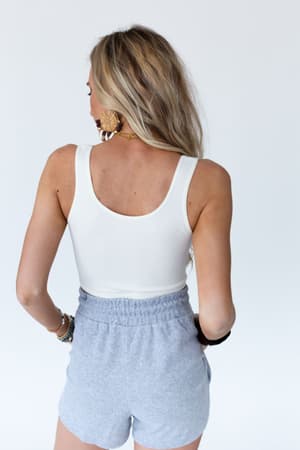 A woman with long blonde hair stands facing away, wearing the Hook It Up Ribbed Brami in ivory and light gray high-waisted shorts. She accessorizes with large earrings and bracelets against a plain white background.