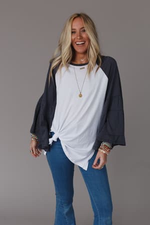 A smiling woman with long blonde hair wears the Bell Sleeve Baseball Tee - Charcoal, tied at the front, paired with blue jeans, layered bracelets, and a necklace. She stands against a plain gray background.