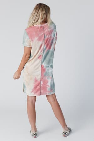 A person with long blonde hair stands facing away, wearing The Nest On The Daily Oversized Slit Tee Dress in a pink, blue, and beige tie-dye print with checkerboard slip-on shoes against a plain light background.