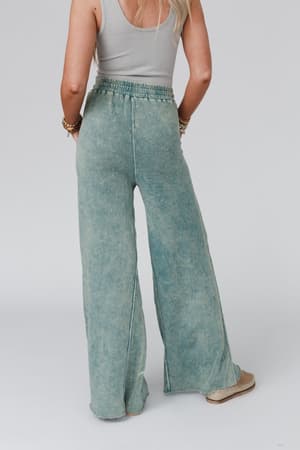 A woman with long blonde hair wears a sleeveless ribbed gray top and the Relaxing Robin Wide Leg Pant in New Teal, standing with her back to the camera against a plain background.