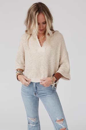 A woman with long blonde hair wears the Just Right Slouchy Sweater in Oatmeal and light blue ripped jeans, standing hands-in-pockets. She accessorizes with bracelets and rings against a plain background.