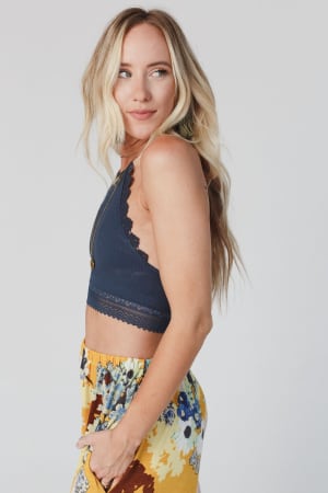A woman with long blonde hair poses in profile, wearing the Light Of The Moon Crochet Trim Bralette in blue gray and colorful floral pants, standing against a plain light background.