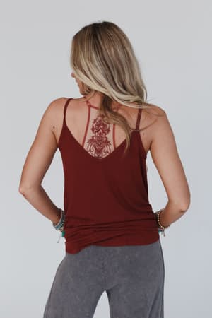 A woman with blonde hair stands with her back to the camera, wearing gray pants and the Tattoo Bralette in Rust, its detailed red back tattoo design visible through the bralette-style straps. She accessorizes with bracelets on both wrists.