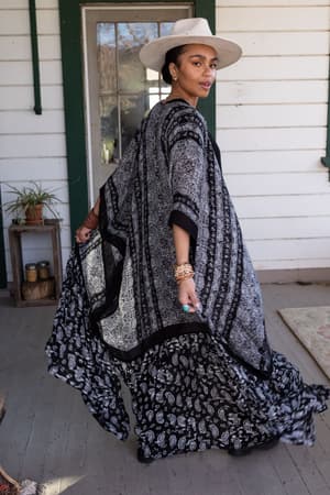 A woman stands on a porch with rustic decor and plants, wearing the Paisley Tapestry Free Flow Duster Kimono in Black and a wide-brimmed hat, turning to look over her shoulder.