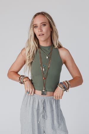 A woman with long blonde hair wears the High Neck Racerback Brami in Olive and light gray pants, accessorized with colorful beaded bracelets and layered necklaces, posing confidently against a plain white background.