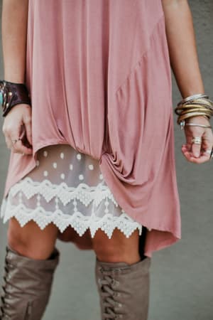 A person in a pink dress with a gathered hem reveals the Layering Love Lace Slip - Ivory underneath. They accessorize with multiple bracelets, rings, and tall taupe boots, all set against a neutral background.