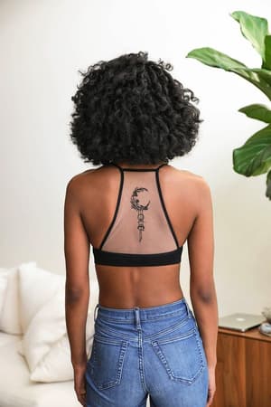 A woman with curly hair wears the Crescent Moon Tattoo Bralette - Black and blue jeans, showing off a crescent moon and key tattoo on her upper back. She stands with her back to the camera; a plant and couch are in the background.