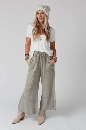A woman stands against a plain background wearing a white knit beanie, white T-shirt, the Feeling Good Wide Leg Pant in Faded Sage, beige ankle boots, and multiple bracelets for a relaxed boho style, with her hands in her pockets.