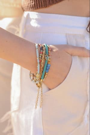 A person in white denim pants displays the Sunburst Stacked Bracelet Set - Multi, featuring colorful beads and gold accents on their wrist, with their hand in the pocket. The scene is captured in soft natural light.