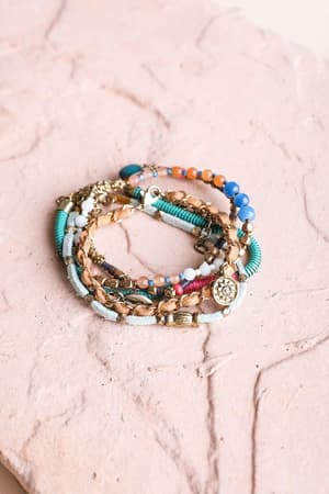 The Sunburst Stacked Bracelet Set - Multi features vibrant, multi-colored boho bracelets with assorted charms, arranged in a circular stack on a textured light pink surface.