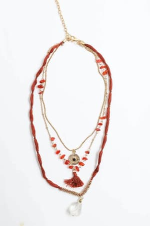 The Carnelian Multi Layered Necklace - Rust features three gold chains with red beads, a central eye-shaped charm with a red tassel, and a large clear teardrop pendant, shown elegantly against a white background.