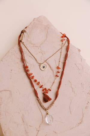 Three layered necklaces on a textured stone: a boho piece with evil eye pendant, one with red beads and tassel, and the Carnelian Multi Layered Necklace - Rust featuring a clear teardrop crystal.