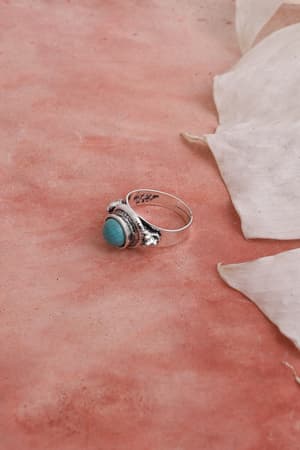 The Kapalua Bay Adjustable Turquoise Ring - Silver, featuring a round turquoise stone, rests on a textured pink surface beside two translucent white leaves—an elegant piece of boho jewelry.