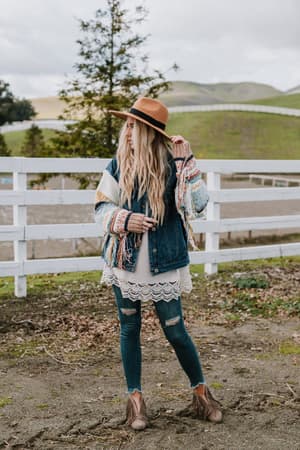 A woman with long blonde hair and a brown hat stands outdoors by a white fence in green hills, wearing the Sukie Sweater Denim Jacket in blue over a lace-trimmed tunic, ripped jeans, and fringe boots.