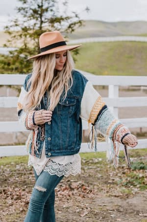 A woman stands outdoors by a white fence, grassy hills behind her, wearing the Sukie Sweater Denim Jacket - Blue with chunky knit sleeves and a wide-brimmed hat, her lace-trimmed top finishing the relaxed bohemian look.