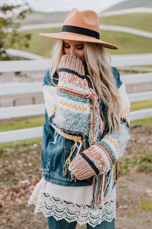 A woman with long blonde hair stands outdoors near a white fence and grassy hills, wearing the Sukie Sweater Denim Jacket - Blue and a tan hat, covering her mouth with her chunky knit sweater sleeve.