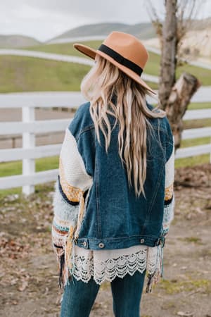 A woman with long blonde hair, wearing a tan hat and the Sukie Sweater Denim Jacket - Blue, stands outdoors facing away near a white fence and grassy hills.
